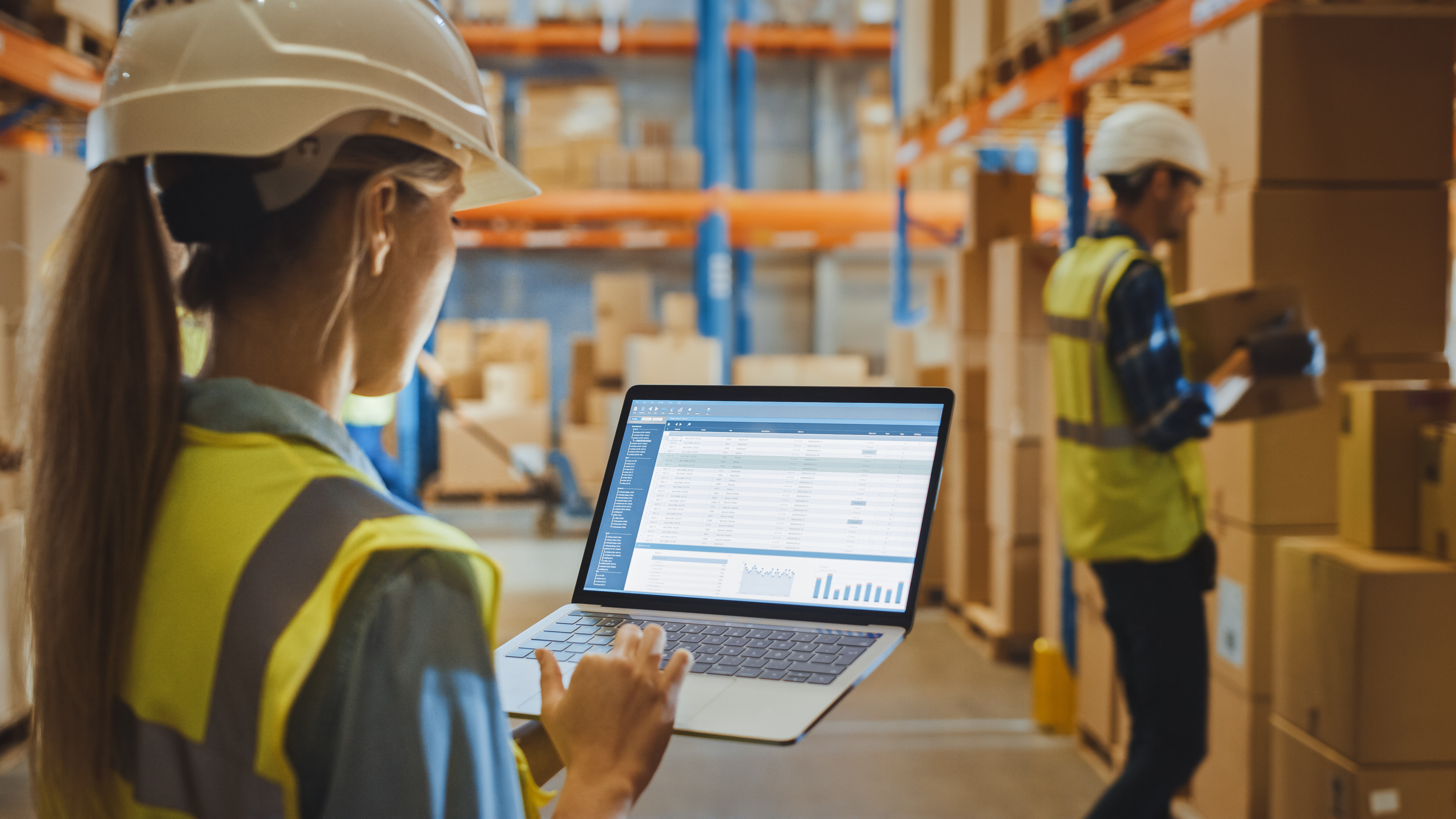 Naispuolinen varastotyöntekijä suojakypärä päässään tarkastelee kannettavalta raporttia, joka liittyy Warehouse Profitability -teemaan.